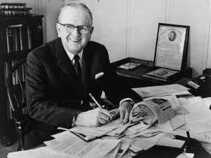 Photo of Norman Vincent Peale smiling working at cluttered office desk.