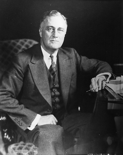 Black and white photo of Franklin Roosevelt sittin at a desk wearing a suit and tie.